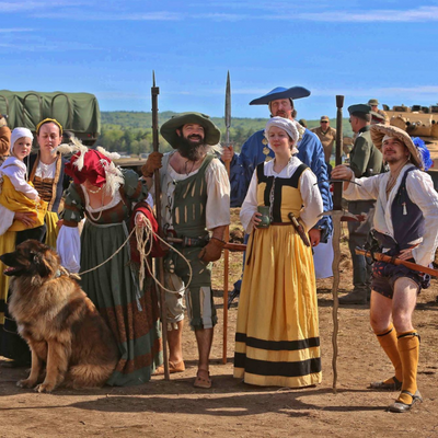 A group of people dressed like it's 1528, with a large dog. There are World War 2 army tanks in the background.