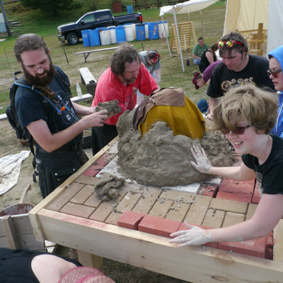 a group of modernly-dressed people standing around a brick-top table, building a cob oven on it.