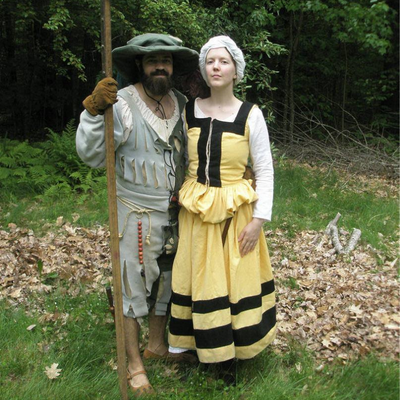 A woman wearing a yellow and black dress standing with a bearded man wearing a blue and grey 1528 outfit and holding a pike.