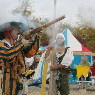 a man wearing a blue and yellow striped outfit fires a matchlock rifle; there is a cloud of smoke and another man behind him.